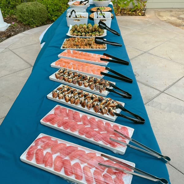 A long table with a blue tablecloth holds various sushi and appetizer platters outdoors.