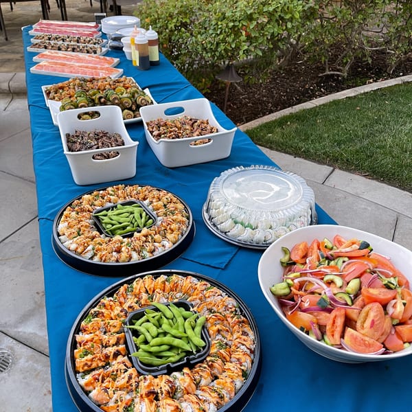 A blue-covered table laden with various sushi platters, salads, and other dishes at an outdoor event.