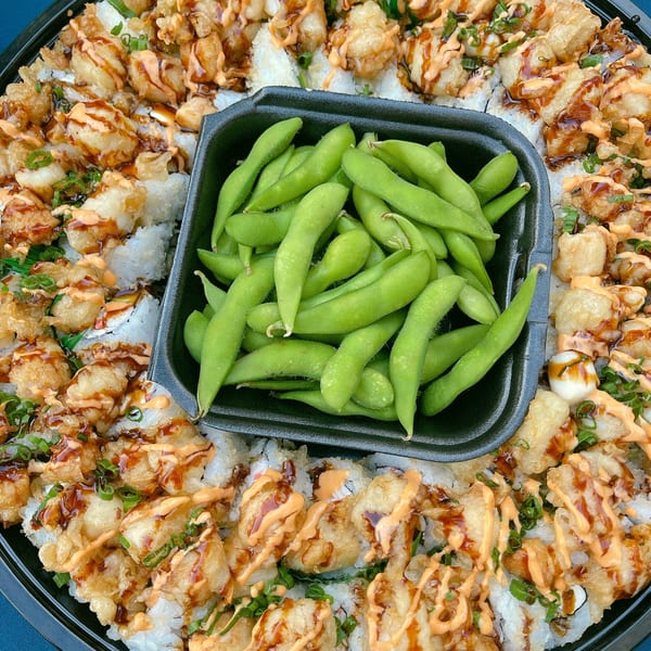 A platter of sushi rolls topped with fried shrimp, sauces, and green onions, with a side of edamame.