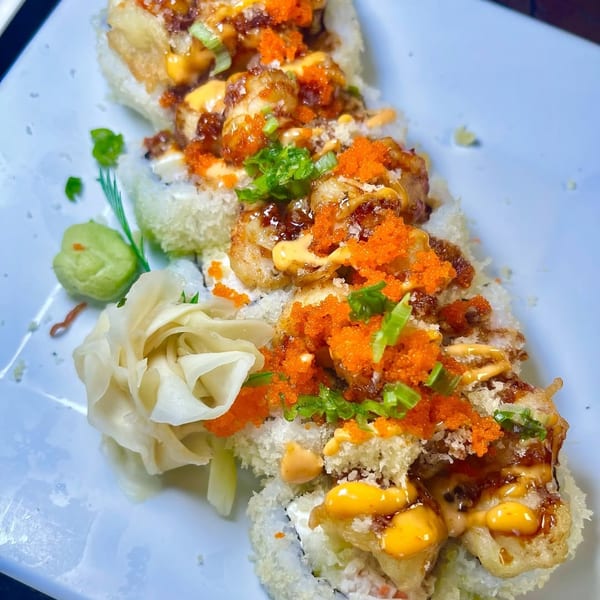 Sushi rolls topped with fried shrimp, orange roe, green onions, and various sauces on a white plate.