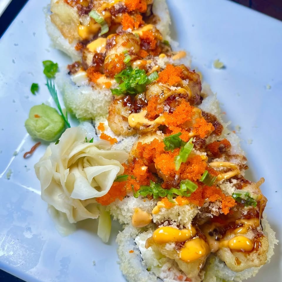 Sushi rolls topped with fried shrimp, orange roe, green onions, and various sauces on a white plate.