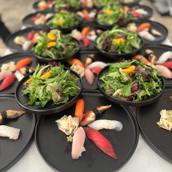 Rows of black plates with sushi and side salads, set for a large gathering.
